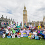 Image shows a group of people with banners in front of the Houses of Parliament