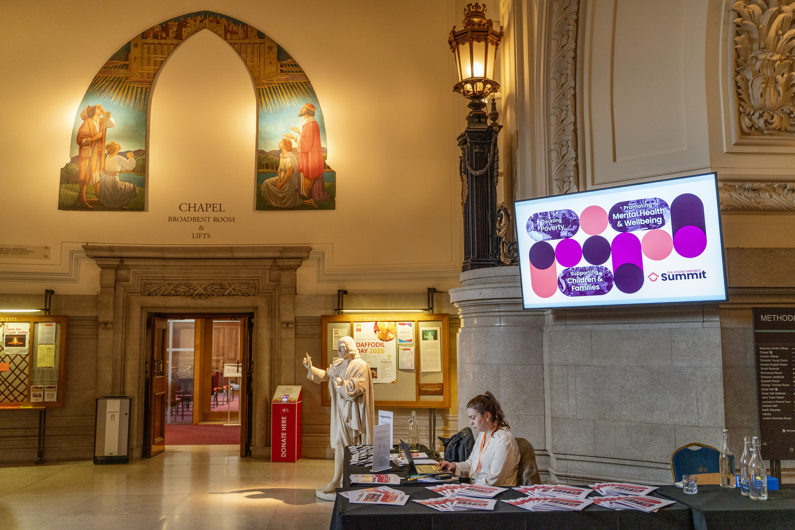 Image shows a woman seated at a table in the foyer of Central Hall in Westminster with ChurchWorks Summit branding on a screen behind her