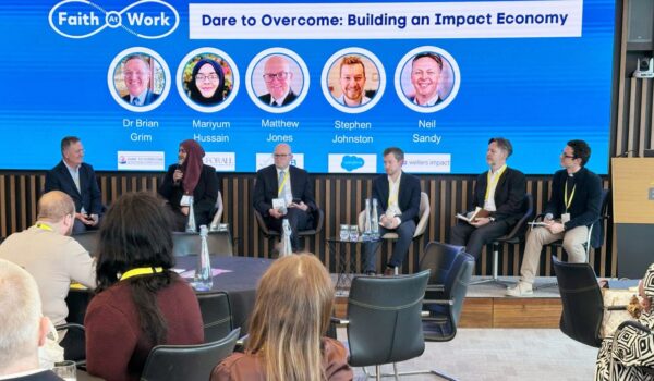 Image shows a group of people seated on a platform with a screen behind them saying Faith At Work Dare to Overcome: Building An Impact Economy