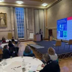 Image shows a person on a stage in front of a large screen presenting to people gathered at tables in a large room