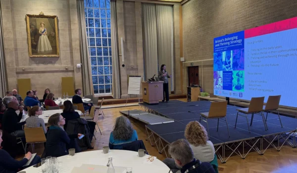 Image shows a person on a stage in front of a large screen presenting to people gathered at tables in a large room