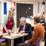 Image shows people seated at tables in a cafe with a smiling woman wearing an apron serving a cup of tea