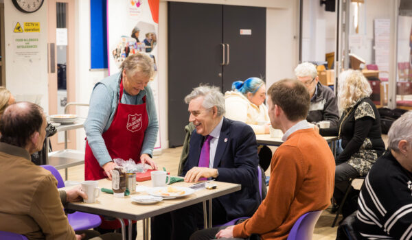 Image shows people seated at tables in a cafe with a smiling woman wearing an apron serving a cup of tea